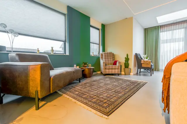 Ruime, lichte woonkamer met een strakke naadloze vloer in warm beige. Grote ramen zorgen voor optimale lichtinval, wanden in groen en geel creëren een verfrist kleuraccent.