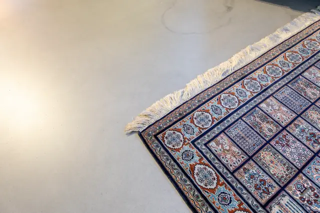 Een minimalistisch ingerichte ruimte met een lichte, naadloze vloer als achtergrond. Een traditioneel Oosters tapijt met rijke kleuren en fijne patronen wordt fraai in beeld gebracht tegen de serene, neutrale ondergrond.