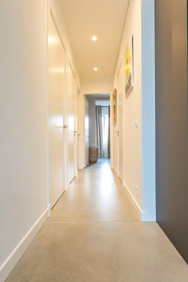 Een sierlijke, lichte gang met perfect afgewerkte naadloze vloer in warm beige en witte wanden. Ingebouwde verlichting creëert een rustige, luxe sfeer met scherpe lijnen en totale ordenlijkheid.