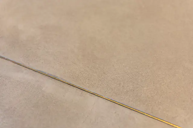 Close-up van een subtiele, grijsbeige naadloze vloer met een smal gouden inlegwerk dat een strakke lijn vormt. Het oppervlak toont een verfijnd, mat afwerking met minimale textuur.
