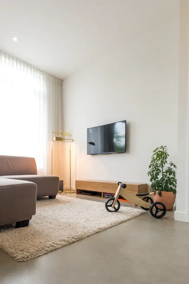 Ruime, minimalistische woonkamer met grote ramen, warme natuurlijke belichting en een naadloze vloer in warme grijs. De ruimte oogt opgeruimd, licht en zeer strak afgewerkt.