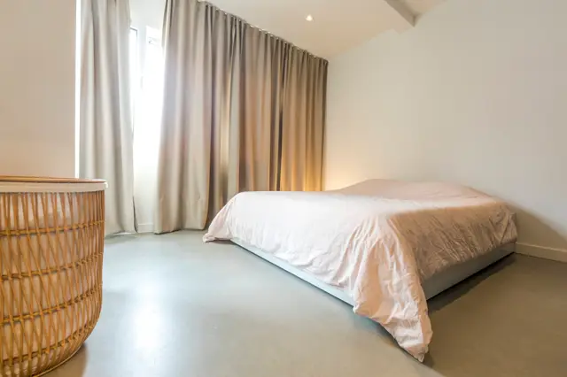 Minimalistisch ingerichte slaapkamer met een grote naadloze vloer in warm grijs, doorlopend onder het bed. Het licht, open design met zachte naturel tonen en hoog plafond creëert een rustige luxe sfeer.