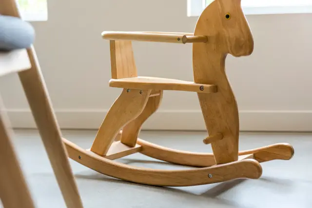 Een fraai houten schommelpaard met natuurlijke afwerking staat op een naadloze lichte vloer. De minimalistische, licht ingerichte ruimte met ramen benadrukt de pure vormgeving van het meubel.