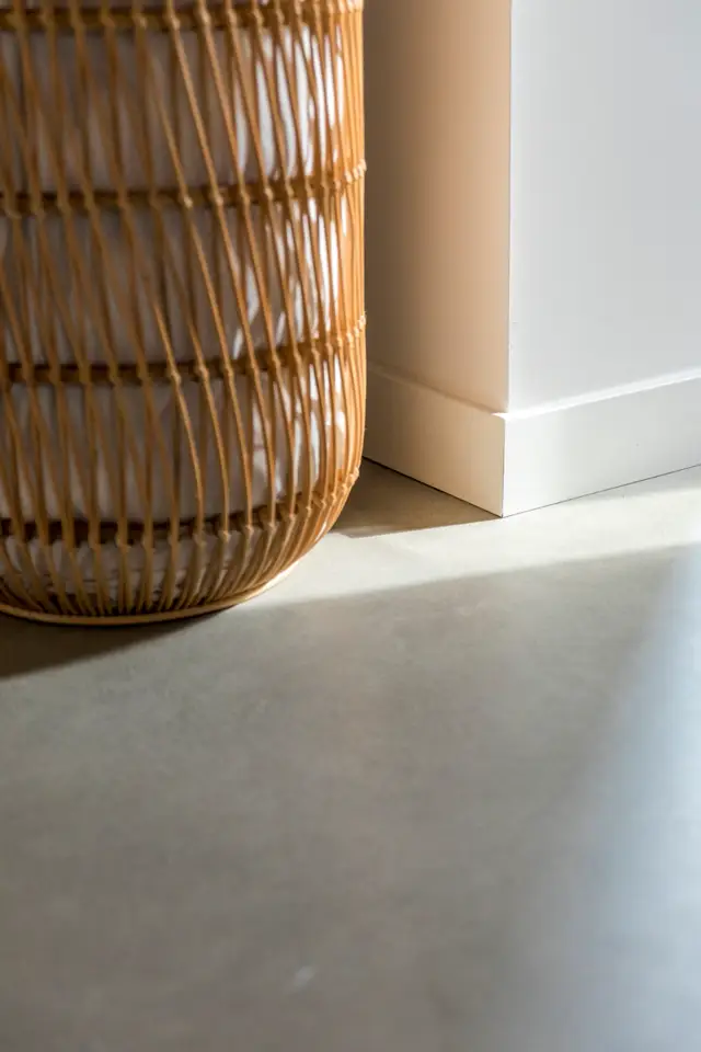 Een minimalistisch interieurdetail met een lichte naadloze vloer, strakke witte wand en organisch gevormde rieten accessoire. Perfect natuurlijke belichting onderstreept de schone lijnen en rustige uitstraling.