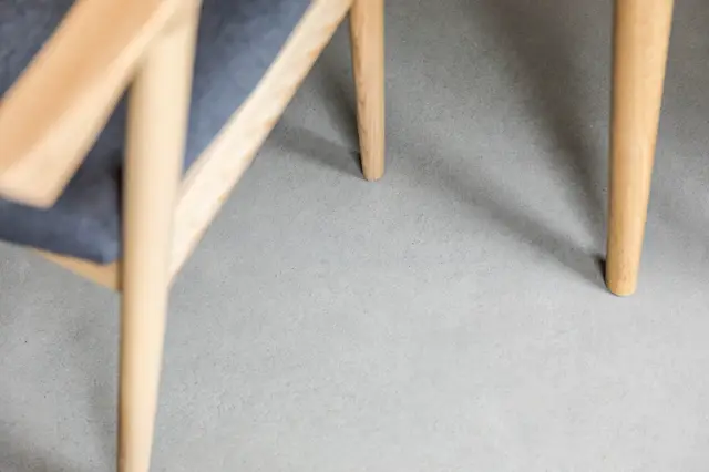 Minimalistisch interieurdetail met een lichte, grijze naadloze vloer en twee houten stoelen met elegante poten. De scherpe schaduwen en warme houtaccenten creëren een serene, hedendaagse sfeer.
