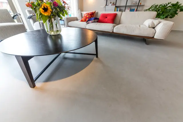 Luxe woonkamer met egale, lichte naadloze vloer die naadloos overgaat in een modern ingerichte ruimte met beige zitmeubel, zwart design tafelwerk en grote ramen met natuurlijk licht.