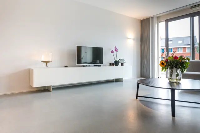 Ruime, lichte woonkamer met een lange naadloze grijs-betonnen vloer die elegant doorloopt. Het interieur straalt rust uit met een wit tv-meubel, minimale inrichting en veel daglicht via grote raampartijen.
