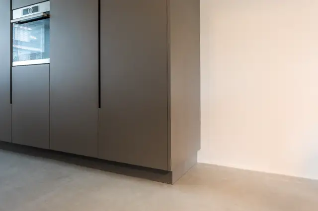Elegant keukenhoek met gesloten kastfronten in warm grijs. De naadloze vloer loopt rustig door onder de keukenblok, scheiding tussen ruimten is subtiel.
