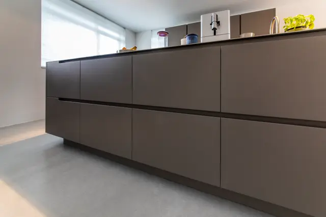 Strak keukeneiland met naadloze fronten in warm grijs-bruin, elegant verlichte open planken boven met minimalistische styling. Heldere daglichtinval, perfecte proporties en schone lijnen.