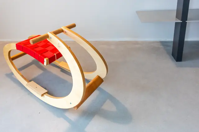 Een minimalistisch houten designobject met rode accenten staat op een lichte, naadloze vloer in een heldere, spaarzaam ingerichte ruimte. De compositie toont zuivere lijnen en perfecte schaduwwerking.