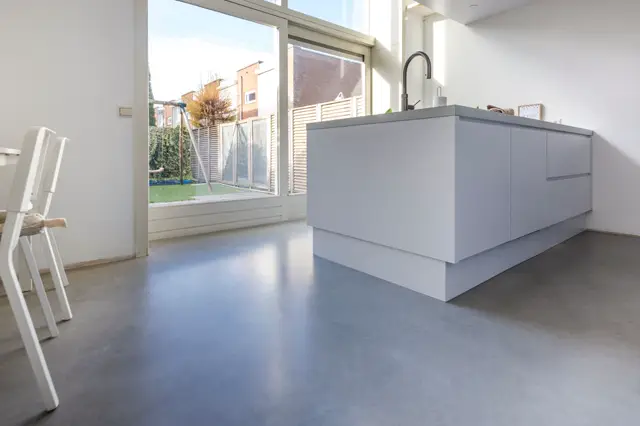 Strakke, moderne keukeneiland in zacht grijs met geïntegreerde kraan, staande op een glanzende grijze naadloze vloer. Grote raampartijen bieden natuurlijk licht en uitzicht op aangelegde tuin.