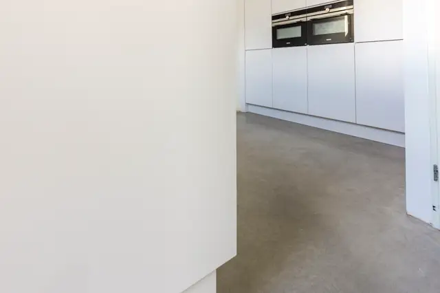Strakke, minimalistische keuken met lichte kasten en geïntegreerde apparatuur. De naadloze vloer in warm grijs loopt doorlopend door de ruimte, wat zorgt voor een geruststellend, opgeruimd effect.