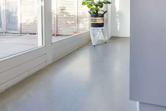 Minimalistisch interieur met een lichte naadloze vloer, grote raampartijen met jaloeziën en een subtiele plantendecoratie. Het ruimtegevoel is open, schoon en aandacht voor detail.