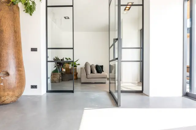 Een licht, minimalistische ruimte met een uniforme grijze naadloze vloer die naadloos doorloopt naar aangrenzende vertrekken. Stalen schuifdeuren met zwarte profielen creëren een strak indelingsgebruik; het interieur is opgeruimd met subtiele groenelementen en neutraal meubilair.
