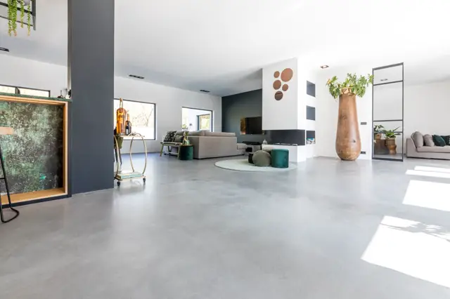 Ruim open woongebied met een strakke, lichte naadloze vloer die de hele ruimte optisch vergroot. Het interieur combineert minimalisme met warme, natuurlijke accenten: houten accessoires, groene plantendecoratie en donkerblauwe wanden als contrast.