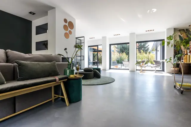 Ruime, licht ingestelde woonkamer met een gepolijste naadloze vloer in grijs-zwart. Open verbinding met terras via grote schuifdeuren, minimalistische inrichting met donkergroene accenten en messing details.