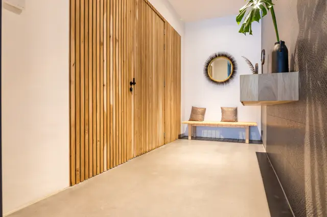 Een serene entreeruimte met een lichte, naadloze vloer die tot de wand doorloopt. Warm houten lamellenwand contrasteert met witte muren, aangevuld met designmeubels en rustieke accessoires.