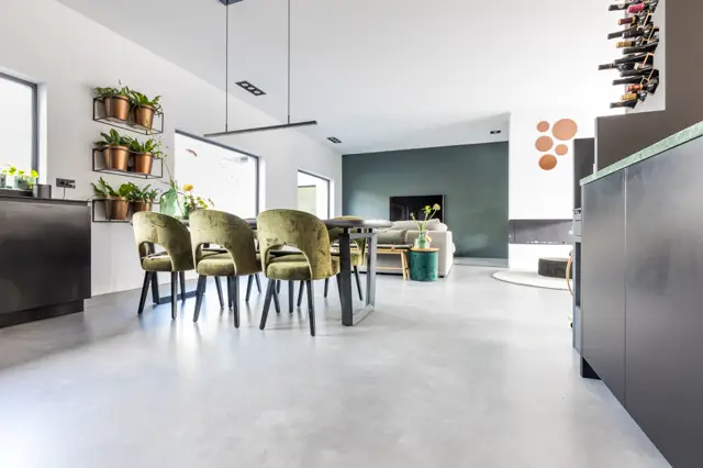 Ruime, lichte woonkeuken met een naadloze lichte vloer die zich naadloos uitstrekt over keuken-, eet- en zitgedeelte. Minimalistisch design met scherpe lijnen, natuurlicht en hoogwaardige afwerking.