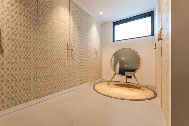 Een serene, licht en ruimtelijk ingerichte kleedkamer met een natuurlijke beige naadloze vloer, textielwanden met geometrisch patroon en elegante gouden accenten. Het natuurlijke licht van het raam accentueert de strakke lijnen en opgeruimde sfeer.