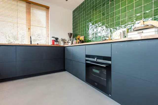 Strakke keuken met donkerblauwe kasten, houten werkblad en opvallende groene tegelwand. De naadloze vloer loopt glad door, aangevuld met natuurlijk licht van houten jaloeziën.