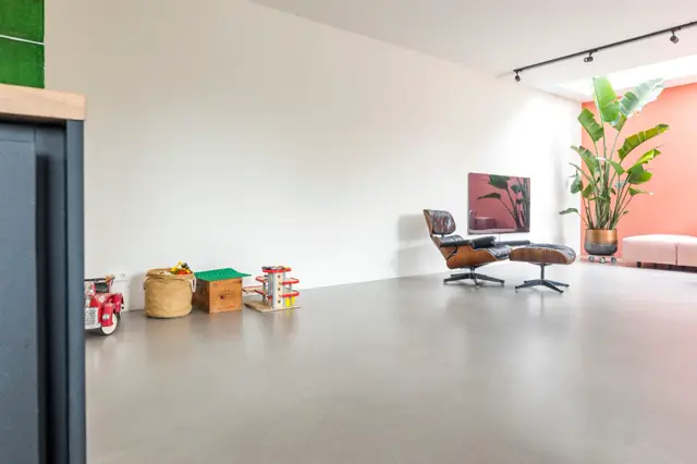 Ruime, lichte woonkamer met een vrijgespeelde naadloze vloer in warm grijs. Het interieur ademt rust en luxe uit met geselecteerde designmeubelen, groenaccenten en zachte roze/coralinvloeden.