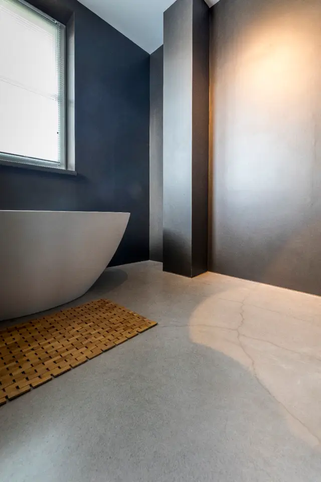 Een strak, modern badkamerinterieur met een elegant vrijstaand bad, donkerblauwe wanden en een lichtvloer die naadloos overgaat. Natuurlijk licht valt door het raam en creëert zachte schaduwspelen.
