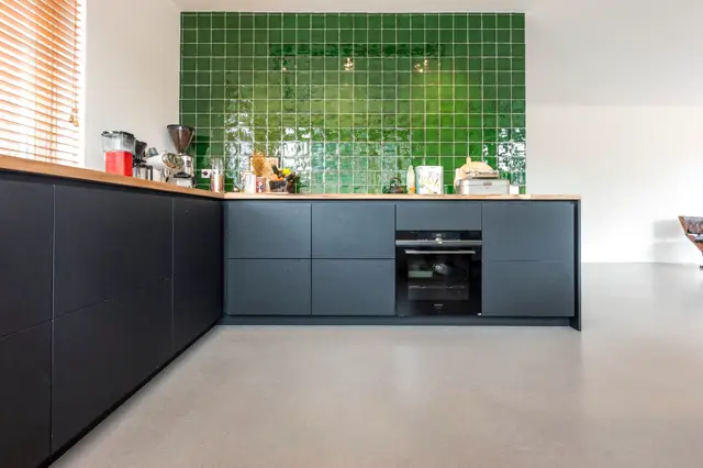 Strakke keuken met donkerblauwe kastenwand, houten werkblad en opvallende groene tegelwand. Naadloze lichte vloer creëert een schoon, minimalistisch geheel.