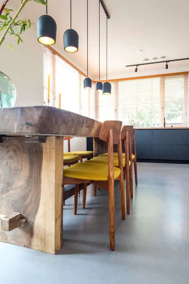 Een moderne eetkamer met een naadloze vloer in neutral grijs, rustieke houten tafel, gele stoelen en zwarte hangende lampen. Grote ramen zorgen voor veel natuurlijk licht.