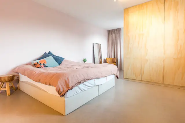 Rustige slaapkamer met een lichte naadloze vloer, witte wanden en natuurlijk houtaccenten. De ruimte ademt minimalisme en elegantie uit met strakke lijnen en een opgeruimde, serene sfeer.