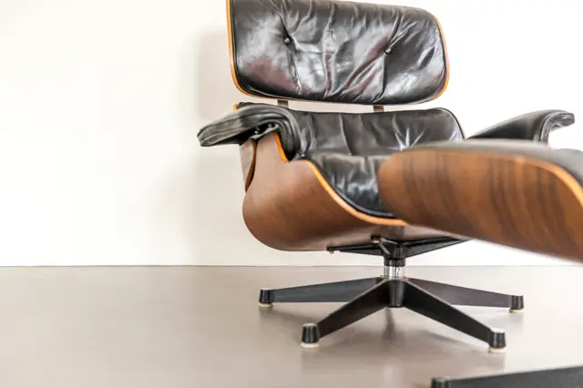 Een iconische leren design stoel met walnoothouten schaal en zwart lederen bekleding staat op een lichte, naadloze vloer. De minimalistische setting met wit muur en glanzende vloer benadrukt het meubel als statement piece.