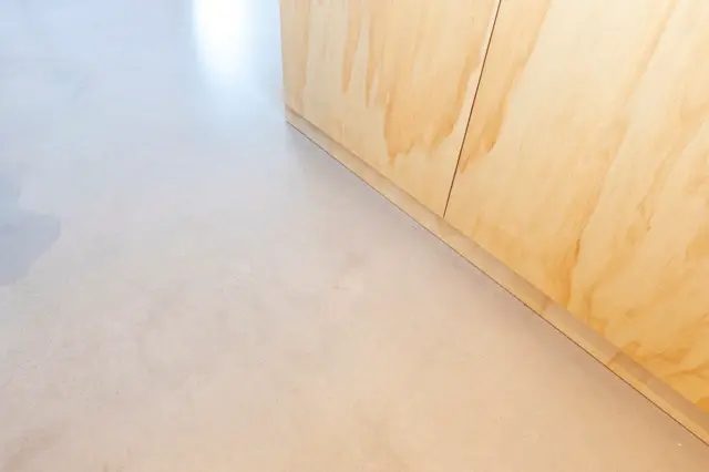 Detail van een lichte naadloze vloer die nauwloos aansluit op een natuurlijk houten kastfront. Het snijvlak is scherp en strak afgewerkt, onderstreept door minimalistisch design.