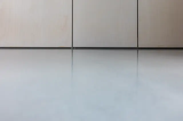 Een strak gecomponeerde ruimte met naadloze wand- en vloeroppervlakken in warme grijstinten. Fijne horizontale lijnen en metallische voegen creëren een serene, ordelijke uitstraling.