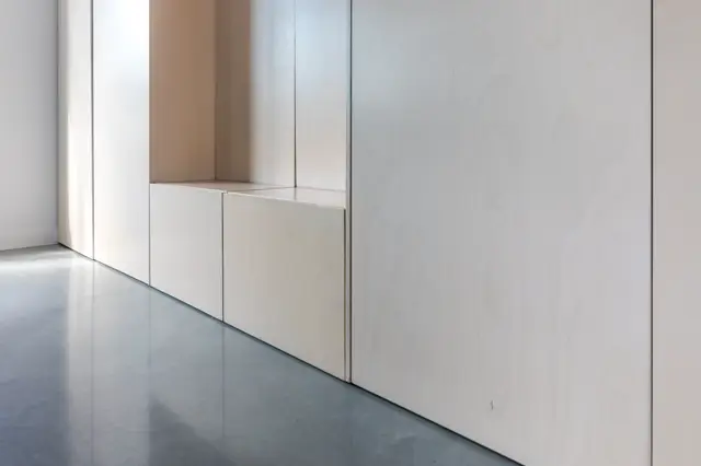 Een strakke, lichte hal met perfecte naadloze wanden in wit en beige tinten. De vloer glans subtiel en creëert een serene, opgeruimde uitstraling met scherpe lijnen en minimalistische architectuur.