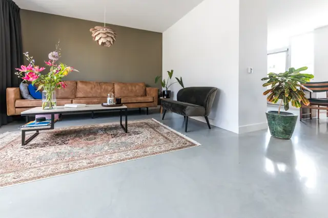 Ruime, lichte woonkamer met glanzende naadloze vloer in grijs. Strak vormgegeven ruimte met cognackleurige sofa, donkere fauteuil en open layout; warm geworden door zachtgroene en roze bloemen in moderne vazen.