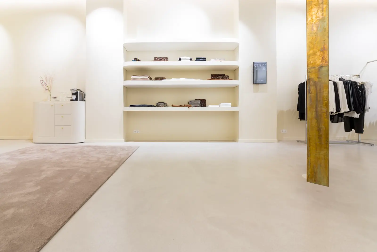 Ruime, lichte bedrijfsruimte met contrast tussen warme en koude naadloze vloerzones. Strakke wandafwerking met ingebouwde kasten en gouden kolom als designelement.