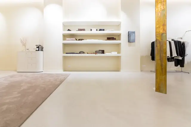Ruime, lichte bedrijfsruimte met contrast tussen warme en koude naadloze vloerzones. Strakke wandafwerking met ingebouwde kasten en gouden kolom als designelement.