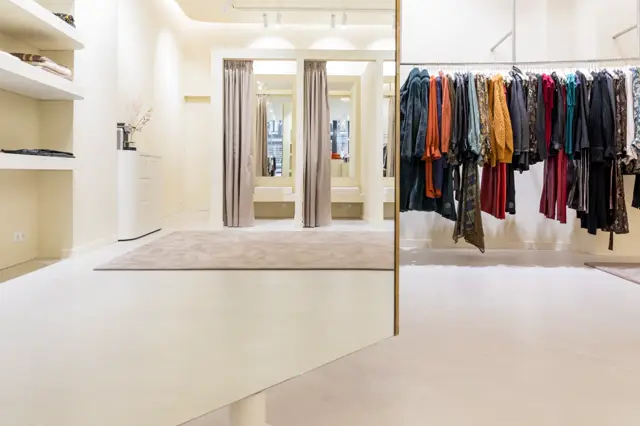 Schoon, licht winkelinterieur met een uniforme naadloze vloer in crème. De ruimte ademt luxe door strakke lijnen, grote spiegels en perfect ingedeeld display met kledingrekken. Natuurlijk licht en subtiele details creëren een verfijnde sfeer.