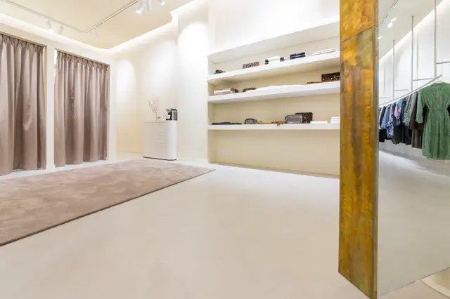 Strakke, lichte garderobe- en aankleedruimte met naadloze vloer in warm beige en twee verschillende zones: een met zacht textiel, een met kledingrek. Volledig opgeruimd en symmetrisch ontworpen.