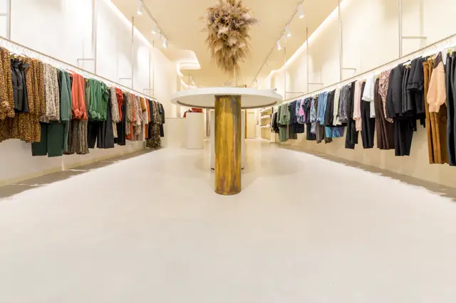 Ruime, lichte winkelruimte met een makeloos witte naadloze vloer die de ruimte optisch vergroten en voor een serene, luxe sfeer zorgt. Het interieur combineert strakke architectuur met warme gouden accenten en droogbloemen.