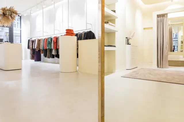 Lichte, minimalistisch ingerichte modewinkel met een naadloze vloer in warm beige. De ruimte ademt rust en elegantie uit dankzij strakke lijnen, perfect uitgevoerde oppervlakken en subtiele gouden accenten.
