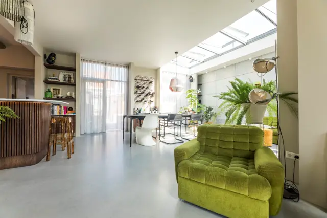 Ruime, lichte woonkamer met naadloze vloer in grijs. Groot raamoppervlak en daklicht zorgen voor optimale lichttoetreding. Strak, minimalistisch interieur met groene accenten en houten details.