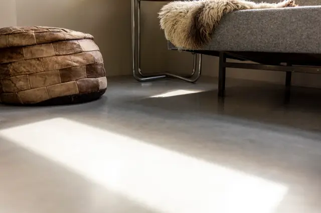 Serene woonruimte met een perfecte naadloze vloer in warm grijs. Natuurlijk licht valt schuin naar binnen en benadrukt de vlakheid en elegantie van de ondergrond. Sprakeloze afwerking met modern meubilair.