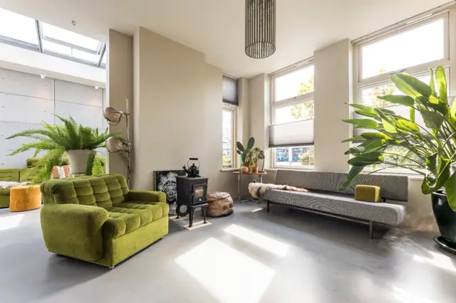 Ruime, lichte woonkamer met grote raampartijen, naadloze grijze vloer en minimalistische inrichting. Frisse accenten met groen en natuurlijke materialen creëren een serene, geordende sfeer.