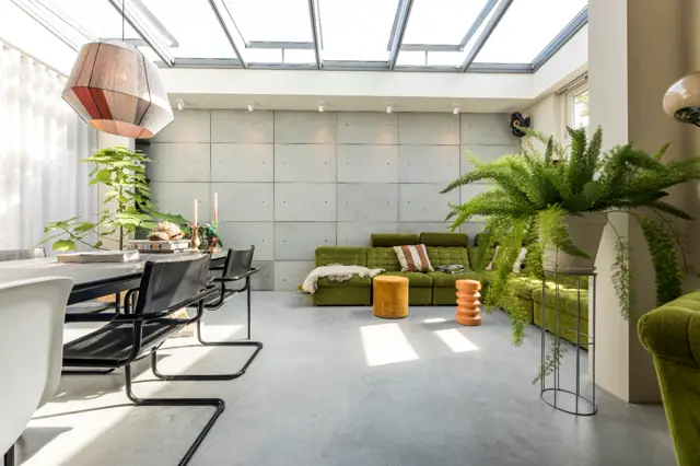 Ruime, lichte woonkamer met grote schuiframen en daklicht. De naadloze vloer loopt ononderbroken door de hele ruimte, onderstreepend de moderne, strakke esthetiek. Minimalistische inrichting met groene accenten.