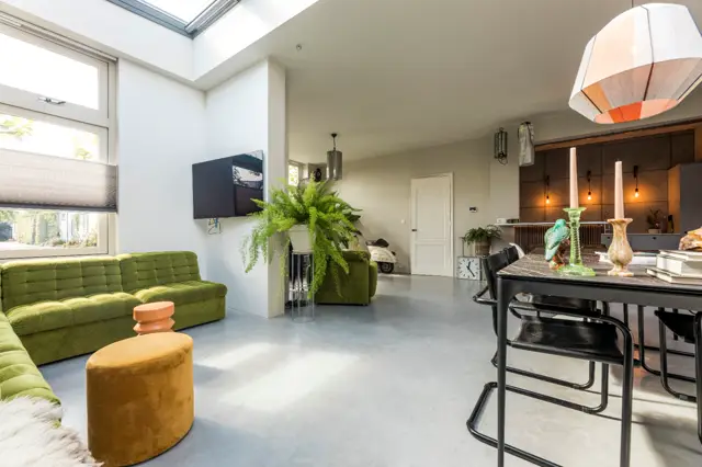 Ruime, moderne woonkamer met een strakke naadloze vloer in licht grijs. De ruimte ademt luxe door grote ramen, natuurlijk licht, minimalistische inrichting en perfect afgewerkte wanden zonder zichtbare voegen.