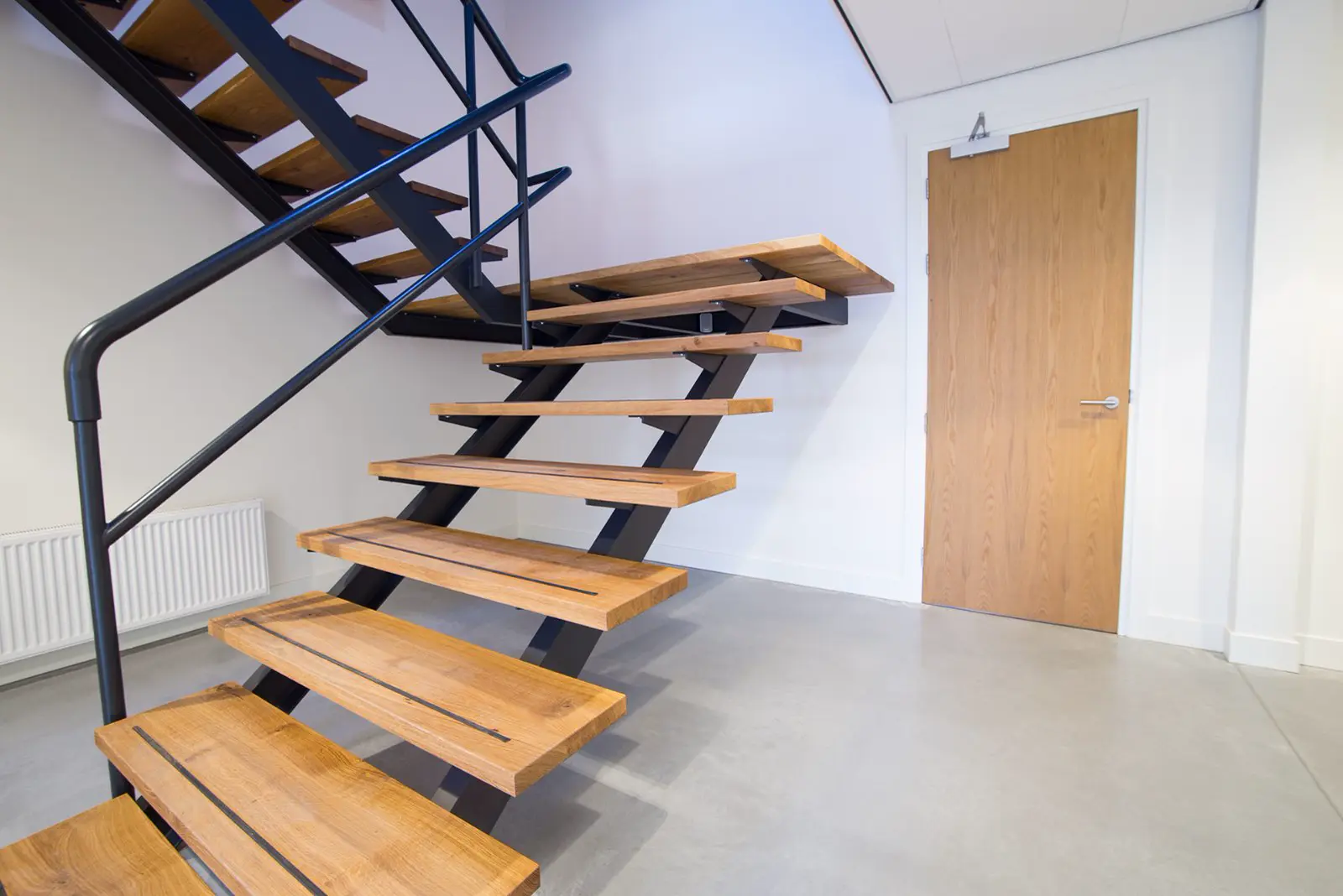 Strakke trap met houten treden en donkerblauwe stalen structuur in een lichte, minimalistisch ingerichte hal met naadloze lichtgrijze vloer.