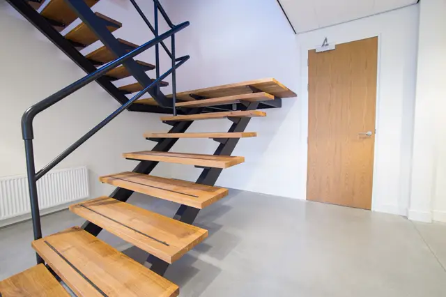 Strakke trap met houten treden en donkerblauwe stalen structuur in een lichte, minimalistisch ingerichte hal met naadloze lichtgrijze vloer.