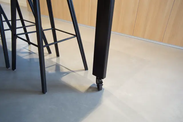 Minimalistische ruimte met glanzende lichte naadloze vloer, zwarte design stoelen en houten wandpanelen. Helder natuurlijk licht creëert scherpe schaduwen en benadrukt de perfecte afwerking.