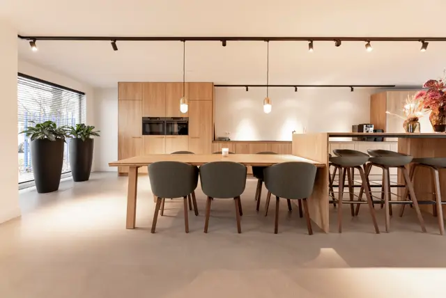Ruim, minimalistisch interieur met natuurlijk licht, houten keukenfronten en een naadloze vloer in warm beige. Strakke lijnen, donkerhouten stoelen en moderne hangende verlichting creëren een luxe sfeer.