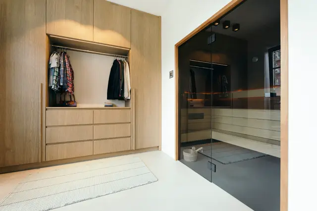 Een strak ontworpen garderobe met eiken houtwerk en open kledingruimte, aangrenzend aan een moderne saunaruimte met glazen deur. Schone lijnen, neutrale kleuren en perfect onderhouden ruimte.